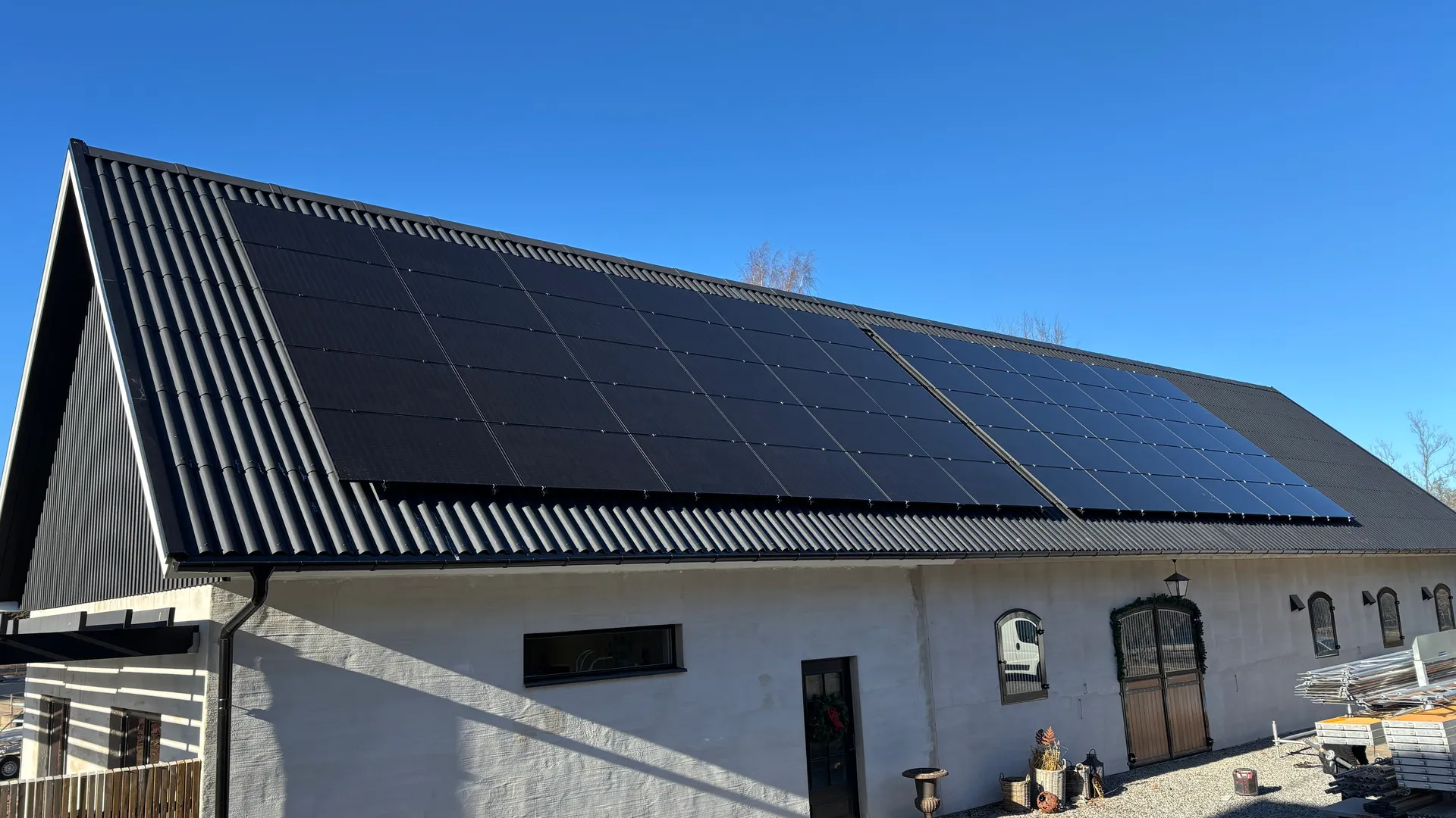 Solceller på ett svart plåttak på en ladugård med vit fasad och blå himmel